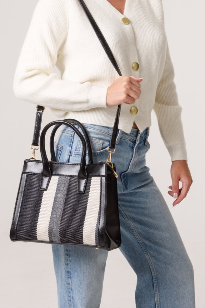 Person holding a black handbag with white and gray stripes, wearing a white cardigan and blue jeans.
