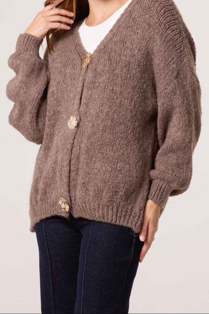 Person wearing a brown knitted cardigan with gold buttons on a plain background